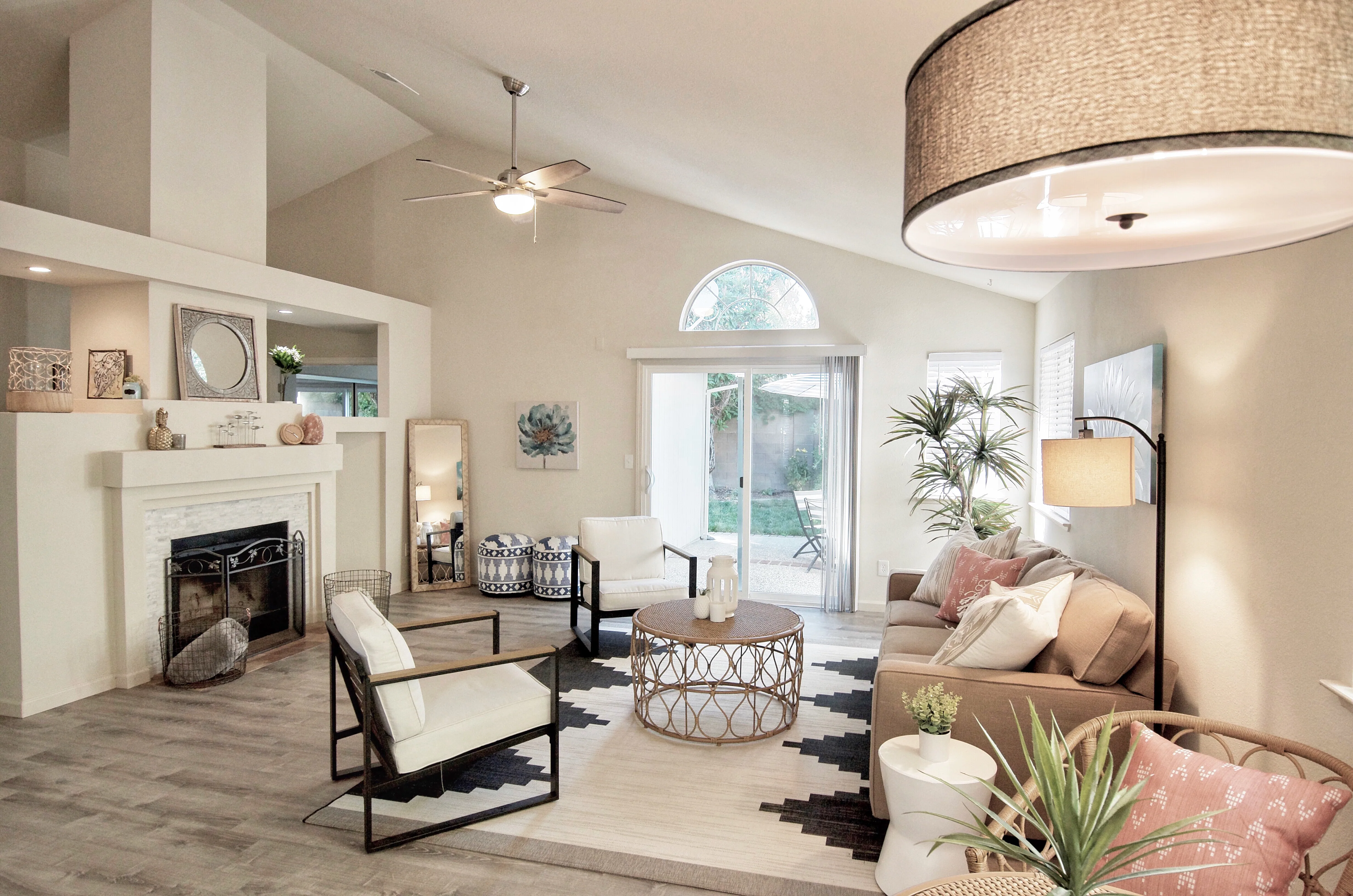 Modern living room with fireplace, cream upholstered chairs, beige sofa, patterned round coffee table, potted plants, and glass doors opening to patio