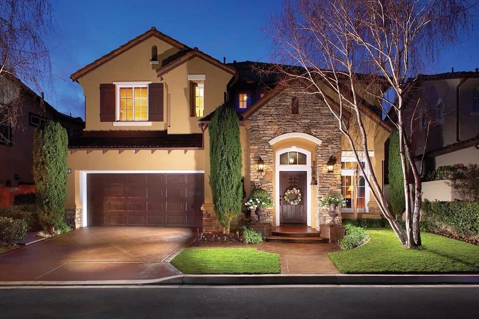 Two-story Mediterranean-style house with tan stucco exterior, stone accents, arched entryway, double garage, lit at dusk with manicured lawn and cypress trees
