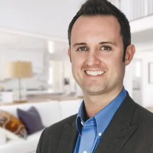Man in blue shirt and dark blazer smiling at camera in front of modern living room