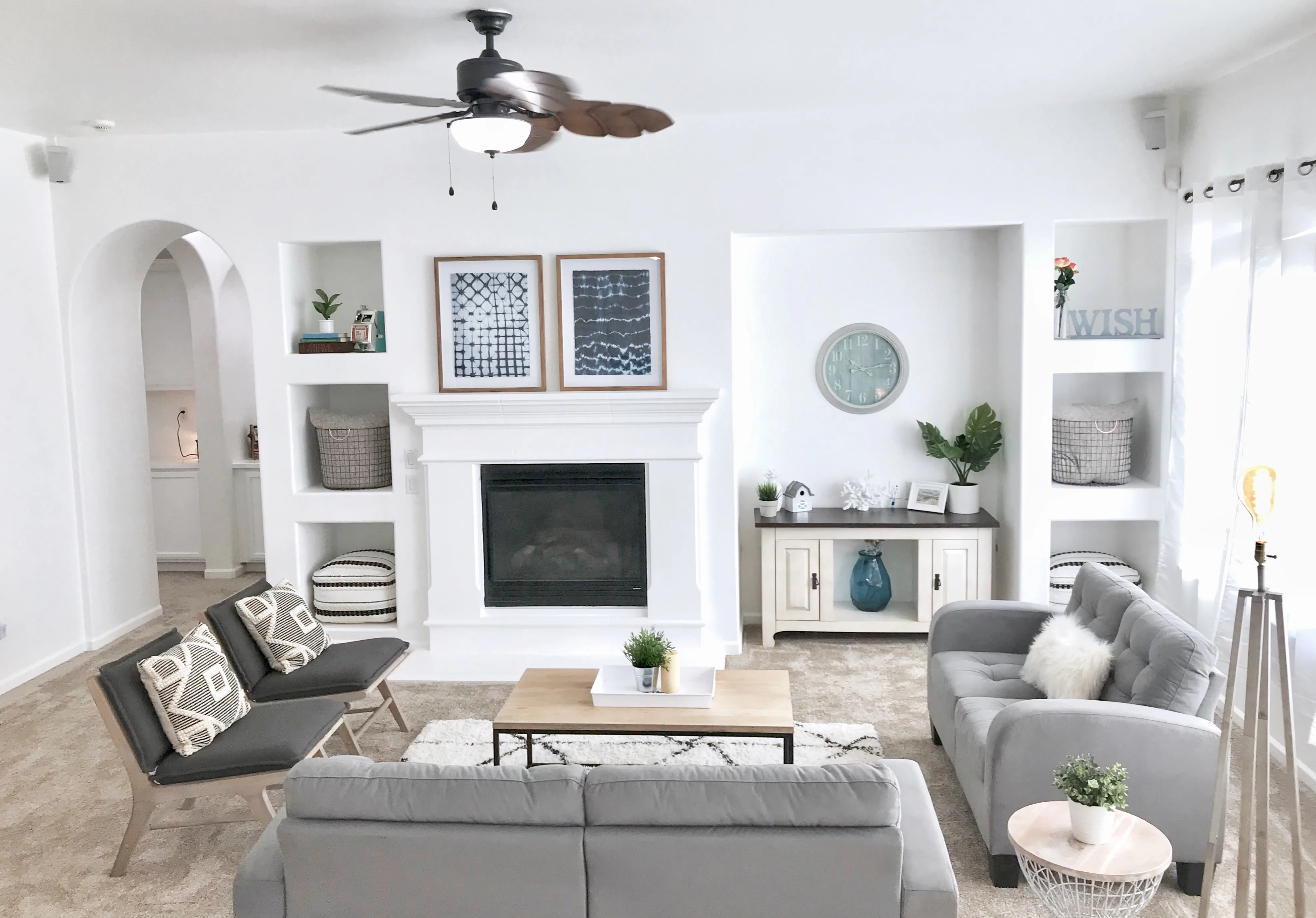 Modern living room with gray and black seating, white fireplace surround, built-in shelving, ceiling fan, and framed artwork