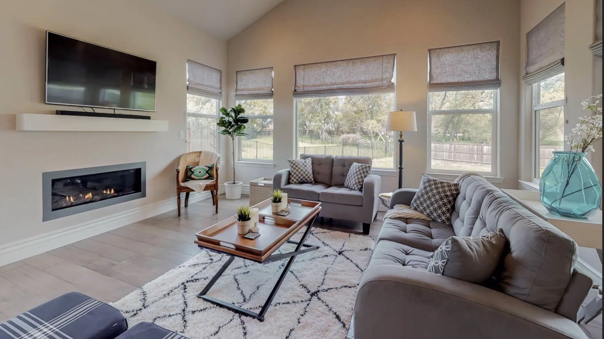 Modern living room with gray seating, fireplace, TV, wooden coffee table, and large windows with natural light