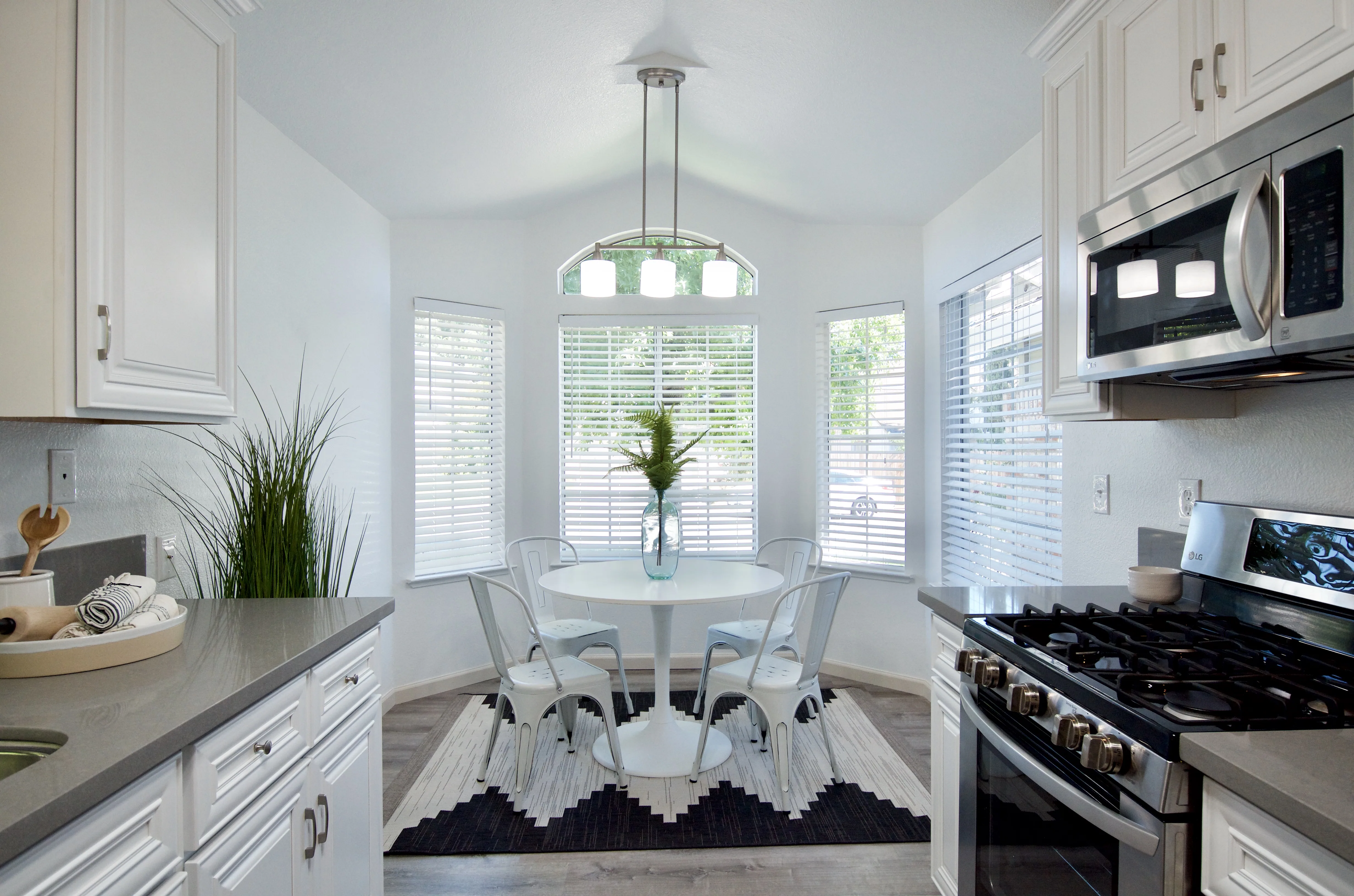 Bright kitchen with white cabinetry, stainless steel appliances, round dining table with chairs by bay windows with plantation shutters, and pendant lighting.
